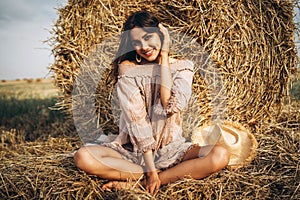 A young woman with long hair and in a dress sits near a hay bale. Woman posing smiling and looking at camera