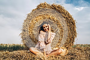 A young woman with long hair and in a dress sits near a hay bale. Woman posing smiling and looking at camera