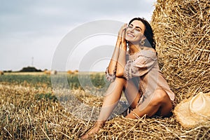 A young woman with long hair and in a dress sits near a hay bale. Woman posing smiling and looking at camera