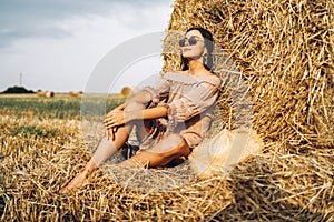 A young woman with long hair and in a dress sits near a hay bale. Woman posing smiling and looking at camera