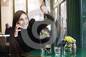 Young woman with long hair gives a hi-five to someone, drink coffee having rest in cafe near windowing