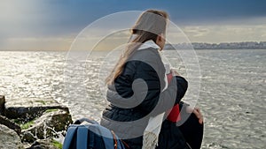 Young woman with long hair sitting on the big rocks at sea coast and looking on stormy weather over the ocean. Concept of tourism