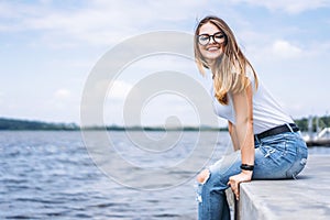 Young woman with long hair in stylish glasses posing on the concrete shore near the lake. Girl dressed in jeans and t-shirt