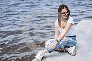 Young woman with long hair in stylish glasses posing on the concrete shore near the lake. Girl dressed in jeans and t-shirt