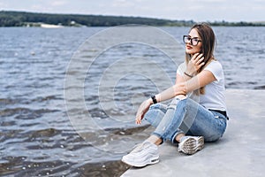 Young woman with long hair in stylish glasses posing on the concrete shore near the lake. Girl dressed in jeans and t-shirt