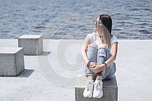 Young woman with long hair in stylish glasses posing on the concrete shore near the lake. Girl dressed in jeans and t-shirt