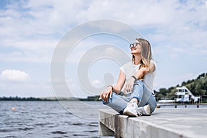 Young woman with long hair in stylish glasses posing on the concrete shore near the lake. Girl dressed in jeans and t-shirt