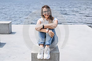 Young woman with long hair in stylish glasses posing on the concrete shore near the lake. Girl dressed in jeans and t-shirt