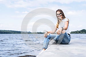 Young woman with long hair in stylish glasses posing on the concrete shore near the lake. Girl dressed in jeans and t-shirt