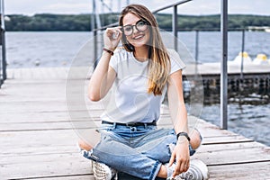 Young woman with long hair in stylish glasses posing on a wooden pier near the lake. Girl dressed in jeans and t-shirt smiling and