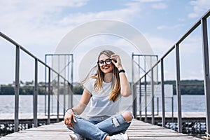 Young woman with long hair in stylish glasses posing on a wooden pier near the lake. Girl dressed in jeans and t-shirt smiling and