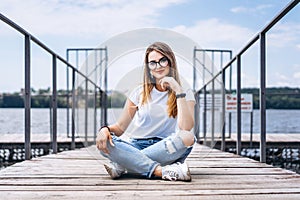 Young woman with long hair in stylish glasses posing on a wooden pier near the lake. Girl dressed in jeans and t-shirt smiling and