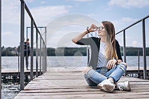 Young woman with long hair in stylish glasses posing on a wooden pier near the lake. Girl dressed in jeans and t-shirt smiling and