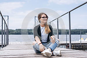 Young woman with long hair in stylish glasses posing on a wooden pier near the lake. Girl dressed in jeans and t-shirt smiling and