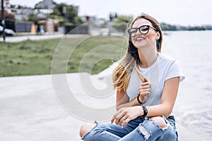Young woman with long hair in stylish glasses sitting on the ground in the city park. Girl dressed in jeans and t-shirt smiling