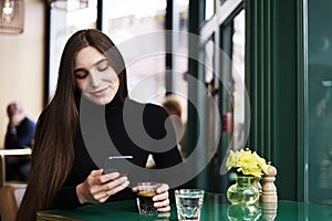 Young woman with long hair texting by mobile phone, smiling, drinking coffee having rest in cafe near window.