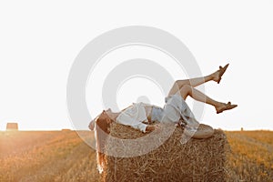 Young woman with long hair, wearing jeans skirt, light shirt is lying on straw bale in field in summer on sunset. Female