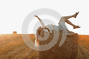 Young woman with long hair, wearing jeans skirt, light shirt is lying on straw bale in field in summer on sunset. Female