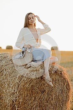 Young woman with long hair, wearing jeans skirt, light shirt and straw bag in hand, sitting on bale on field in summer