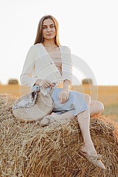 Young woman with long hair, wearing jeans skirt, light shirt and straw bag in hand, sitting on bale on field in summer