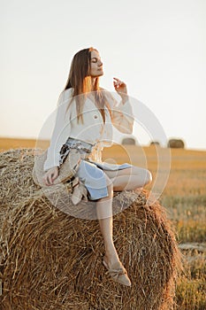 Young woman with long hair, wearing jeans skirt, light shirt and straw bag in hand, sitting on bale on field in summer. Female