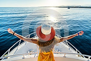 Young woman with long hair wearing yellow dress and straw hat standing with raised hands on white yacht deck enjoying view of blue