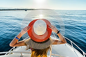 Young woman with long hair wearing yellow dress and straw hat standing on white yacht deck enjoying view of blue sea water