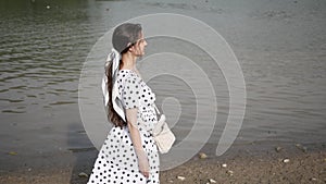 Young woman with long hair in white dress walks along lake with ducks in Park