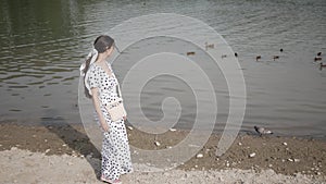 Young woman with long hair in white dress walks along lake with ducks in Park