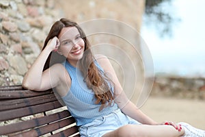 Young woman with long, pretty hair smiles, looking at the camera while resting while sitting on a park bench