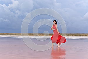 A young woman in a long red dress walks on the lake before the coming storm