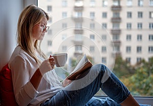 Young woman with long red hair in glasses in a white shirt sitting on the windowsill at home reading a book drinking coffee