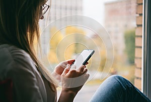 Young woman with long red hair in glasses in a white shirt sitting on the windowsill at home using a smartphone