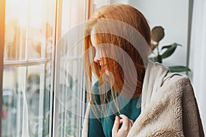 Young woman with long red hair looking through the window at home wrapped in warm comfy blanket