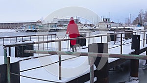 Young woman in long red jacket passes through snow-covered pier and looks into distance at frozen river in winter