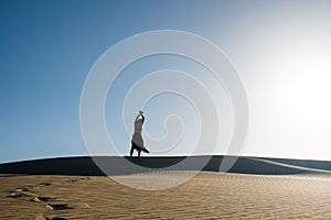Young woman with long skirt dancing in the distance in evocative and confident way on top of desert dune with clear blue sky
