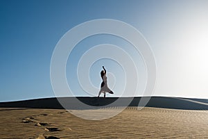 Young woman with long skirt dancing in the distance in evocative and confident way on top of desert dune with clear blue sky