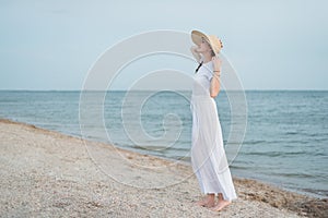 Young woman in long white dress and hat standing on beach. Evening sea