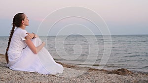 Young woman in long white dress sitting on beach at sunset and looking into distance. Slow motion