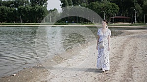 Young woman in long white dress walks along shore of lake