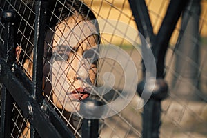 Young woman look through wire fence on sunny day. Woman with fashion look, makeup. Beauty model with makeup face skin