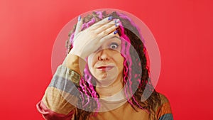 Young woman looking at camera and doing facepalm on red background. Portrait of perplexed female in state of shock