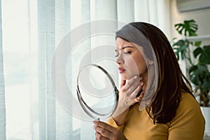 Young woman looking herself in the mirror at home. Worried about acne caused by wearing a mask