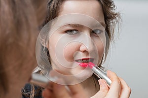 Young woman is looking in mirror and applying red glossy lipstick on her lips