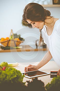 Young woman looking for a new recipe for cooking in a kitchen. Housewife is making online shopping by tablet computer