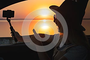 Young woman looking out car window of the sunset on the sea and and photographed on the phone.