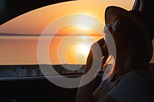 Young woman looking out car window of the sunset on the sea and talking on the phone.