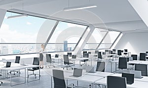 A young woman is looking out the window in the modern bright open space loft office. White tables equipped by modern laptops and b