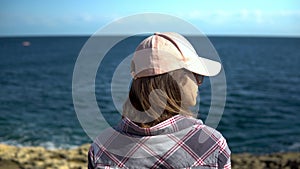 A young woman is looking at the sea. The girl in the cap admires nature. Woman stands with her back to the camera.