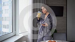 Young woman looking into smartphone while drinking orange juice in the kitchen at home. Woman with healthy food indoors